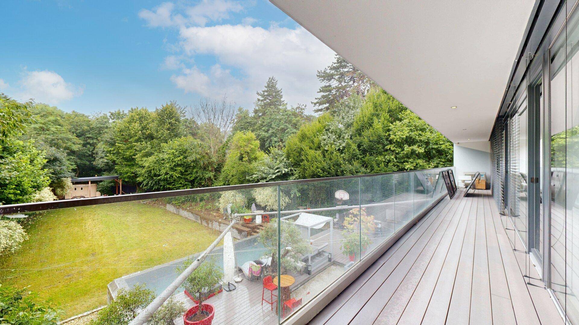 Großer Balkon mit Holzdeck, Glasgeländer und weitem Blick auf den Garten und die Bäume.