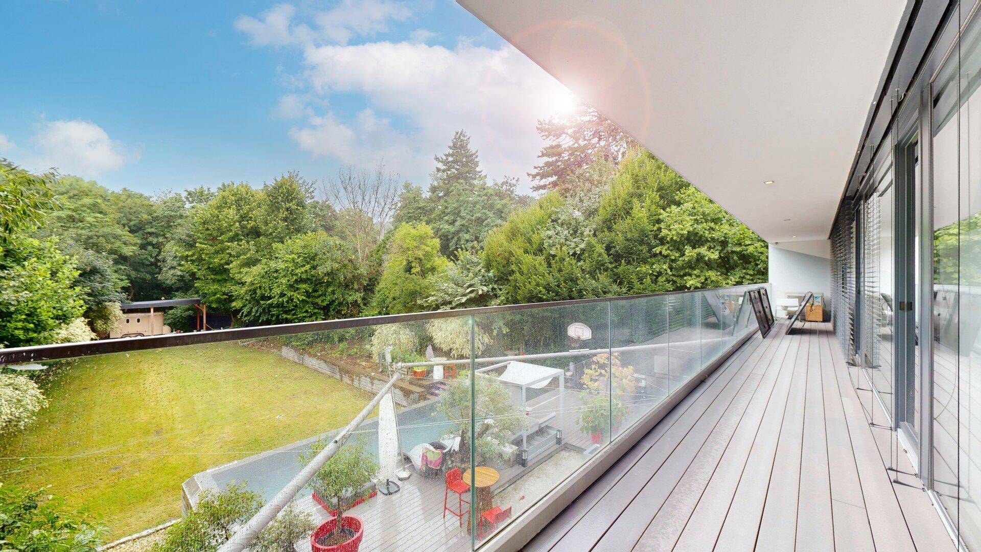 Sonniger Balkon mit Holzdeck, Glasgeländer und Panoramablick auf den grünen Garten.
