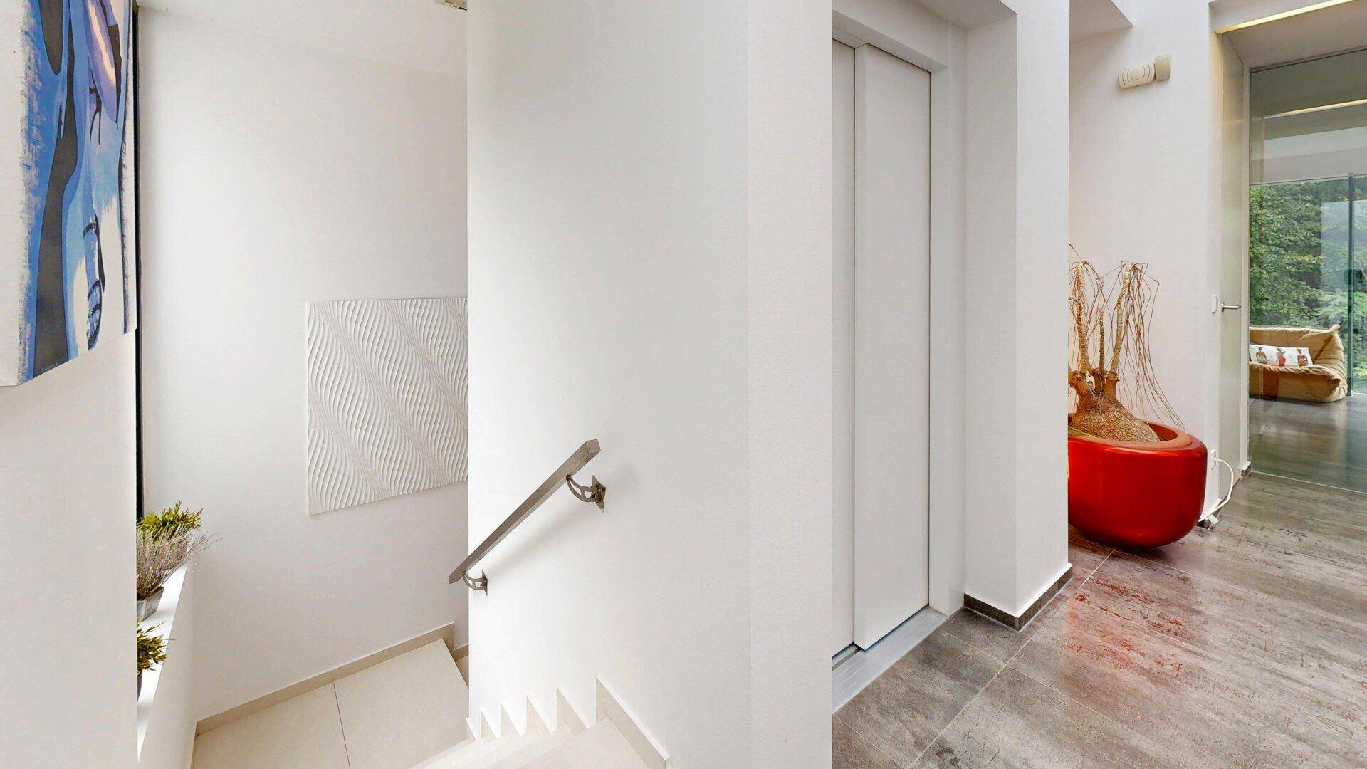 Heller Flur mit Treppe, Handlauf und Aufzug, der verschiedene Ebenen verbindet.