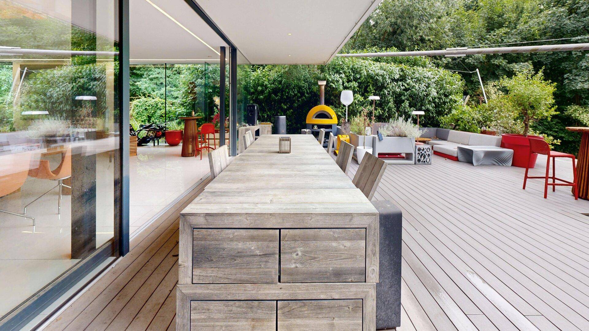 Weitläufige Holzterrasse mit großem Esstisch und gemütlicher Lounge-Ecke im Grünen.