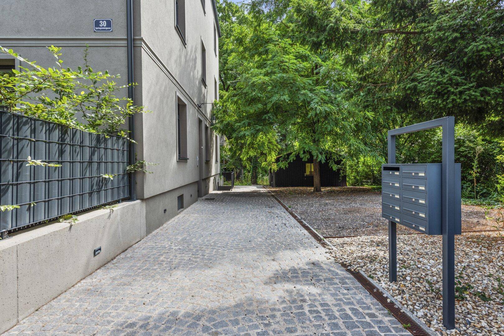 Gepflasterter Weg neben dem Gebäude mit Briefkästen und viel Grün, führt zu einem Nebengebäude.