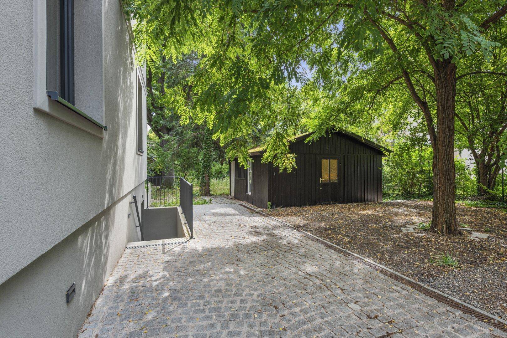 Gepflasterter Weg führt zu einem dunklen Holzschuppen im Grünen, mit Treppe zum Keller.