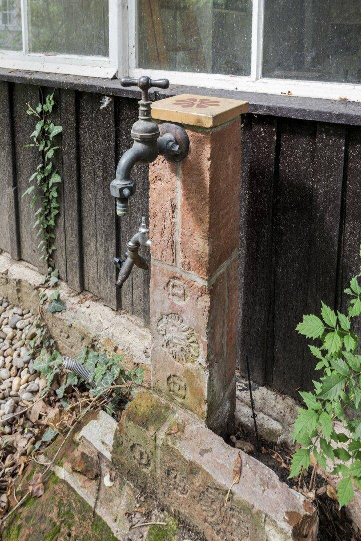 Rustikaler Wasserhahn an einem Steinsockel vor einer Holzwand, umgeben von Kies und Pflanzen.