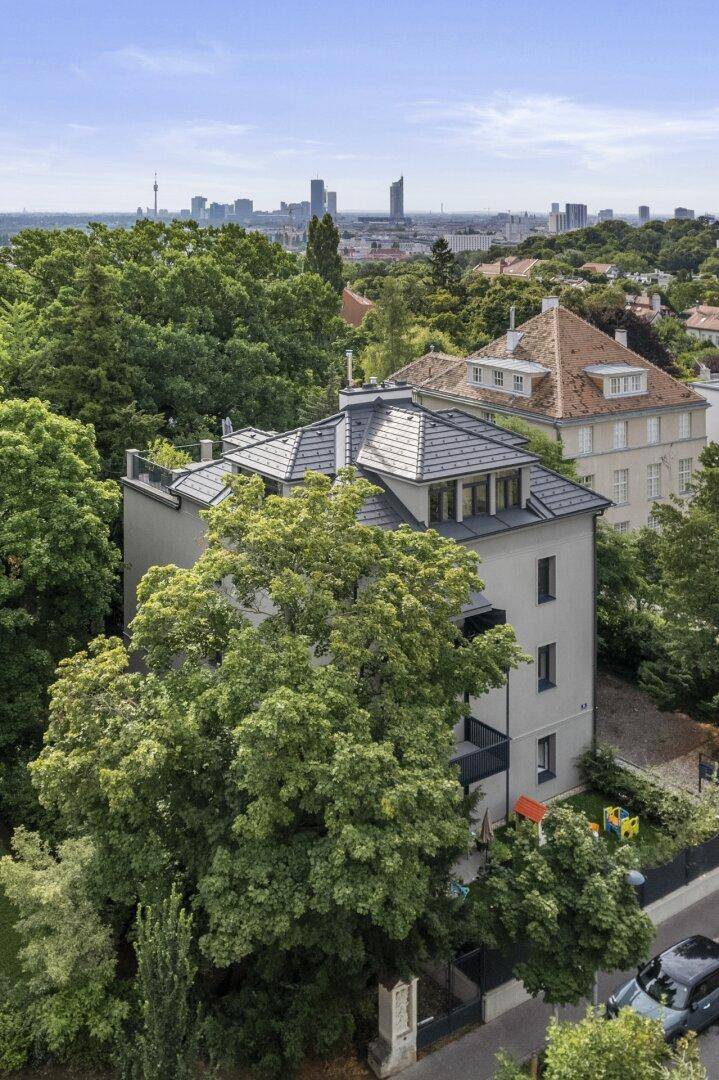 Seitenansicht eines modernen Gebäudes mit Balkonen, umgeben von Bäumen und mit Stadtblick im Hintergrund.