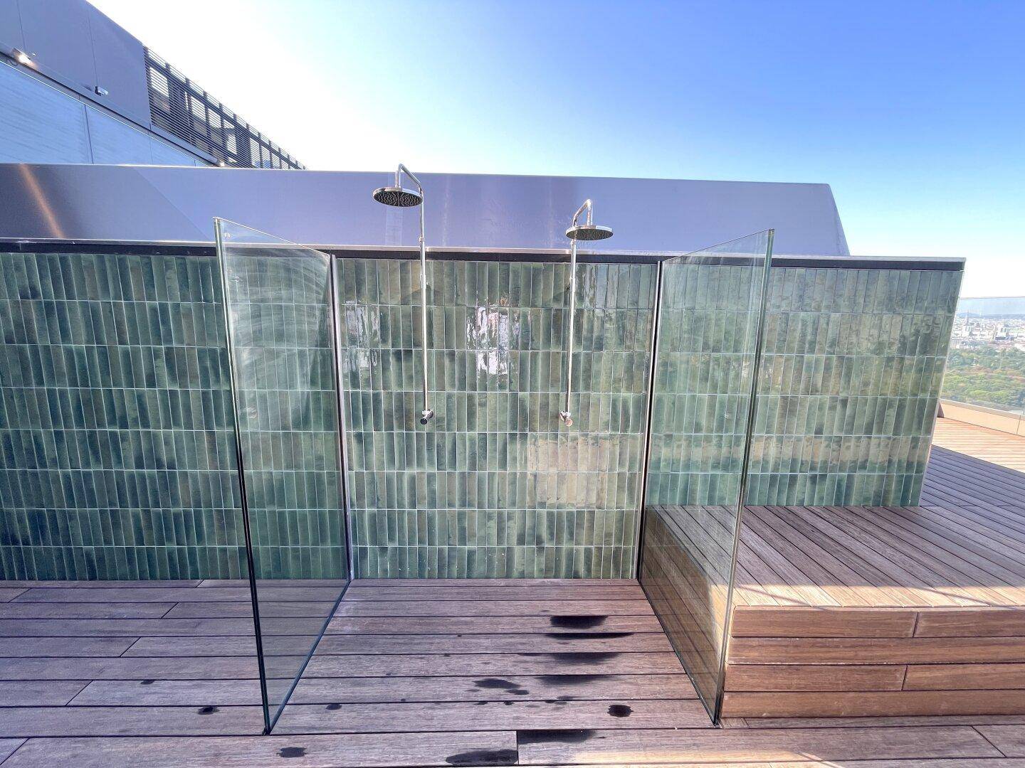 Moderne Außenduschen auf einer Holzterrasse mit grünen Fliesenwänden unter blauem Himmel.