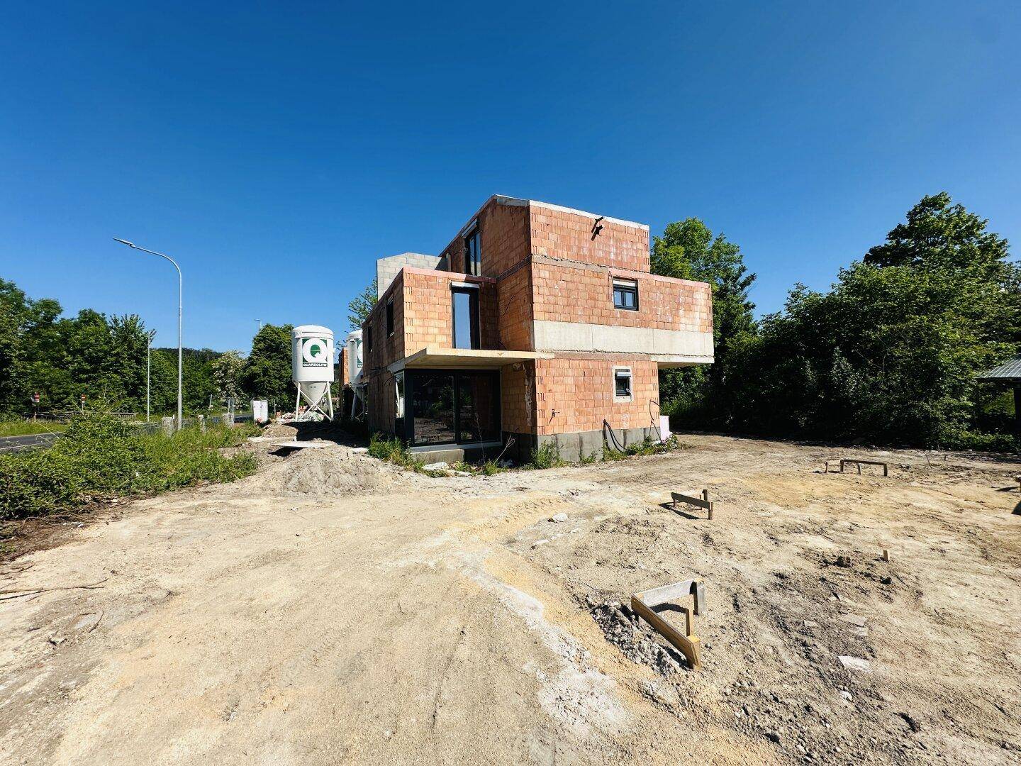 Neubau im Rohbau auf einem weitläufigen Grundstück mit Baustellenelementen und Bäumen im Hintergrund.