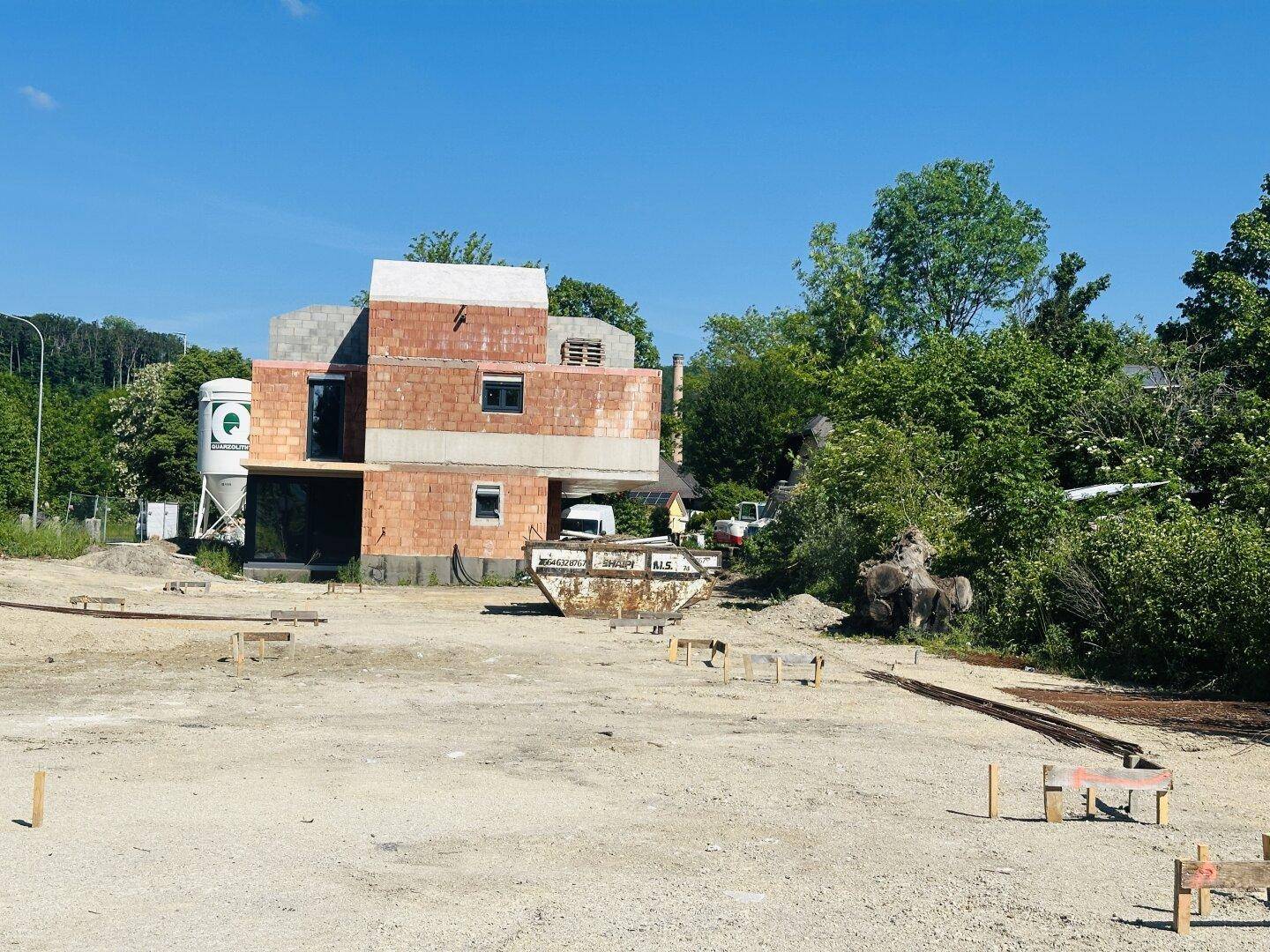 Baugrundstück mit Neubau im Rohbau, Bauschuttcontainern und Bäumen im Hintergrund an einem sonnigen Tag.