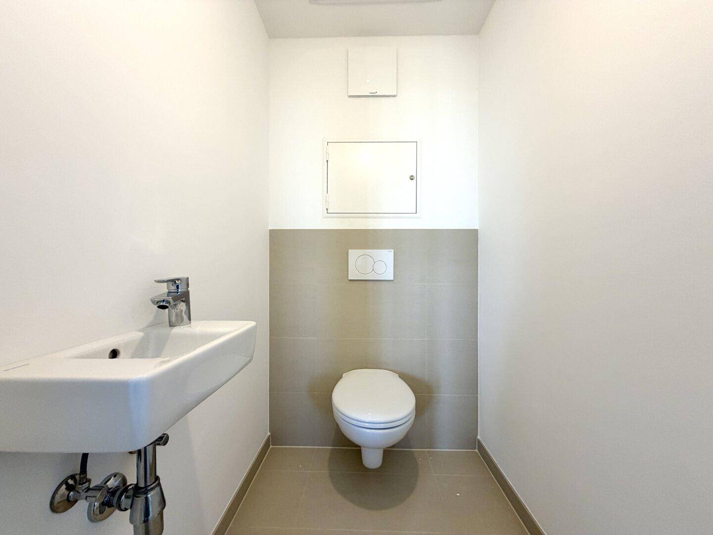 Separates WC mit wandhängender Toilette und kleinem Waschbecken, halbhoch gefliest.