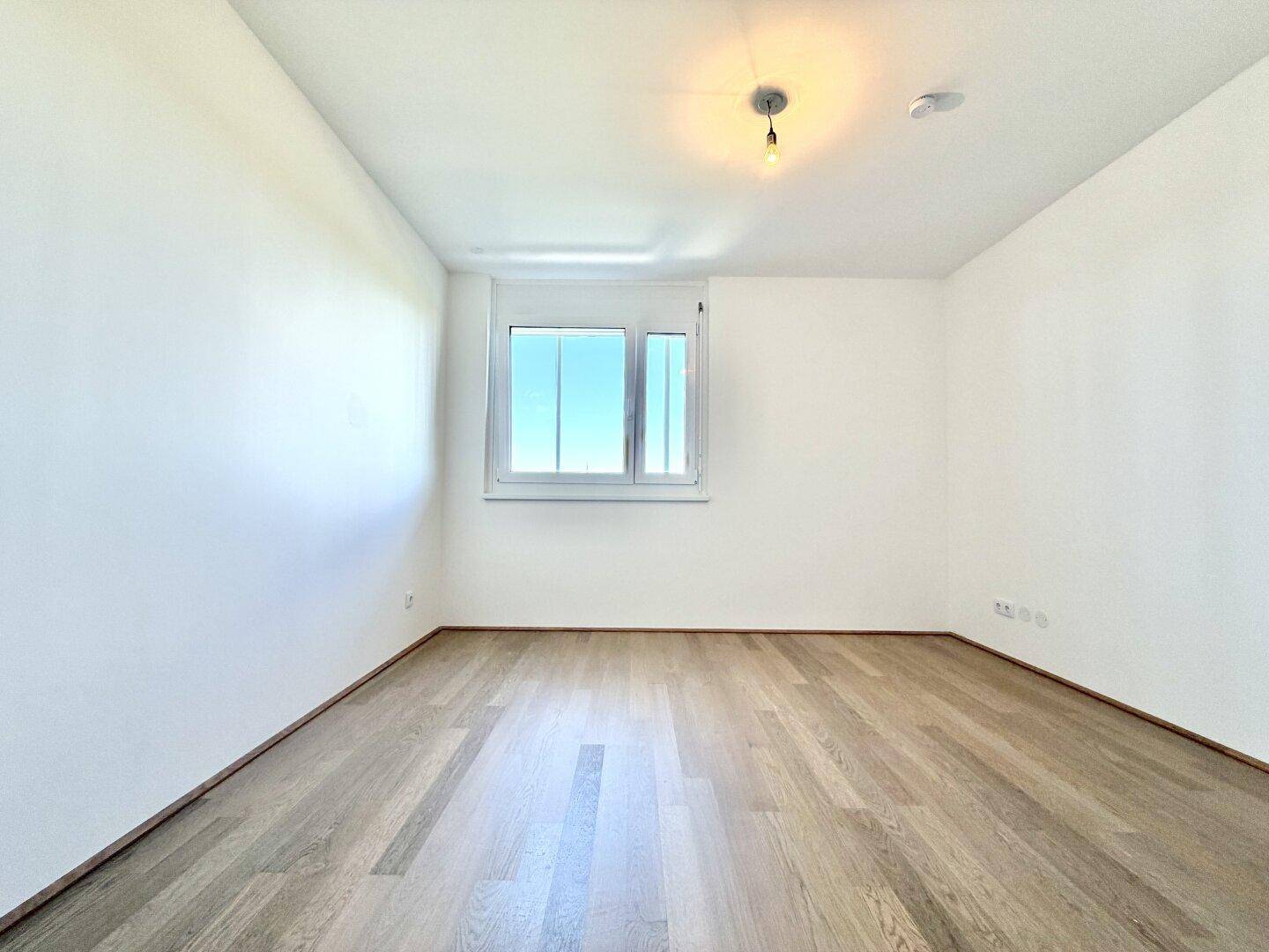 Helles Schlafzimmer mit Parkettboden und einem großen Fenster, das viel Licht spendet.