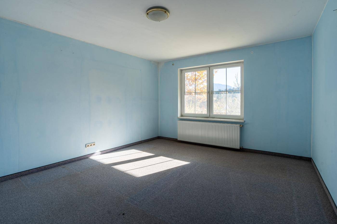 Schlafzimmer mit hellblauen Wänden, Teppichboden und einem Fenster, das viel Tageslicht hereinlässt.