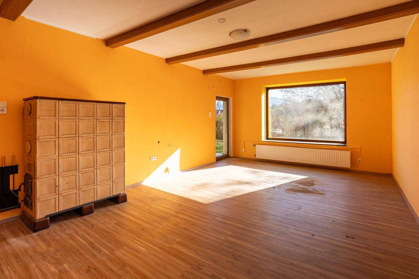 Wohnzimmer mit leuchtend orangefarbenen Wänden, Holzbalken und einem traditionellen Kachelofen.