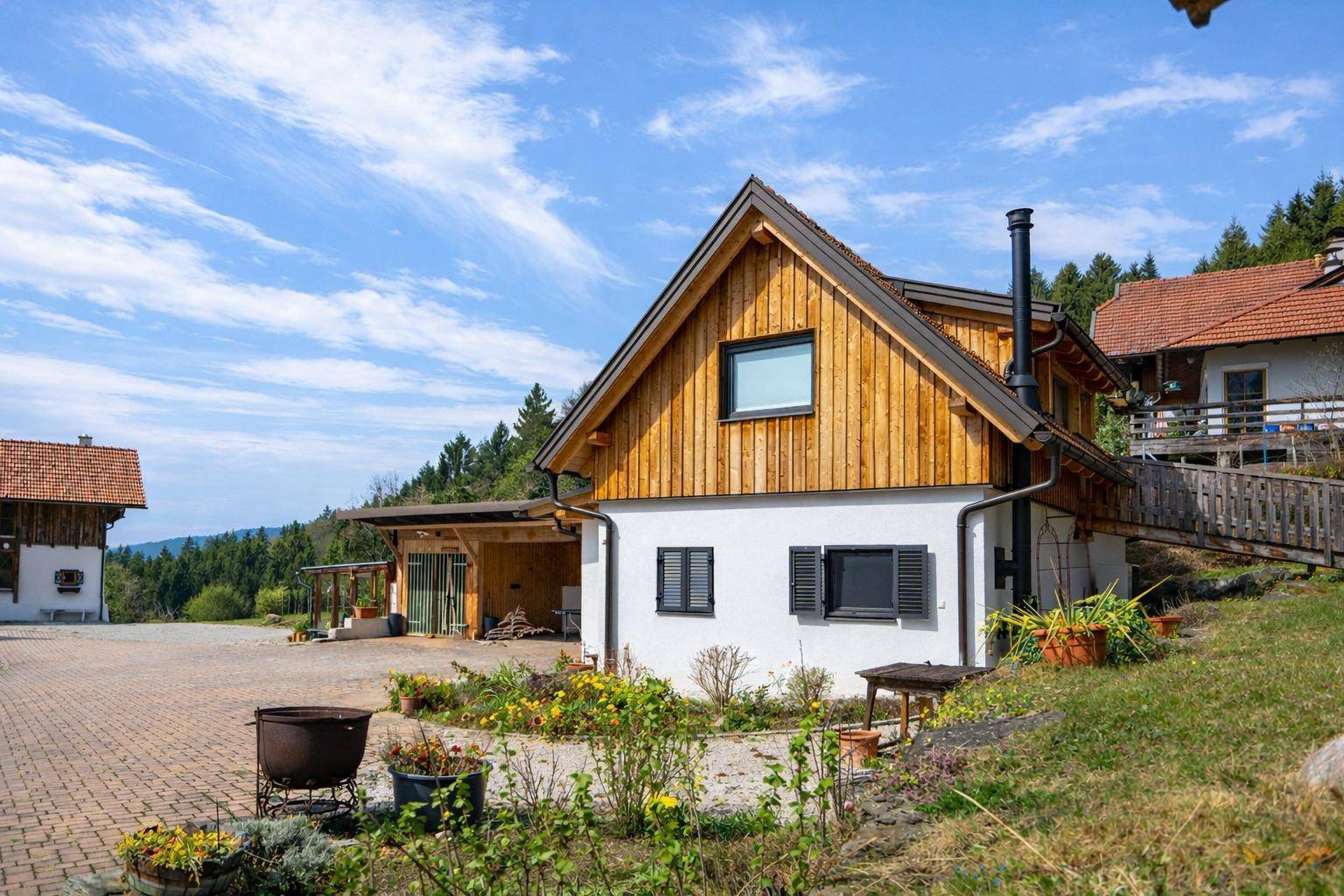 Traditionelles Gebäude mit weißer Fassade, Holzverkleidung und einem Carport, umgeben von Blumenbeeten.