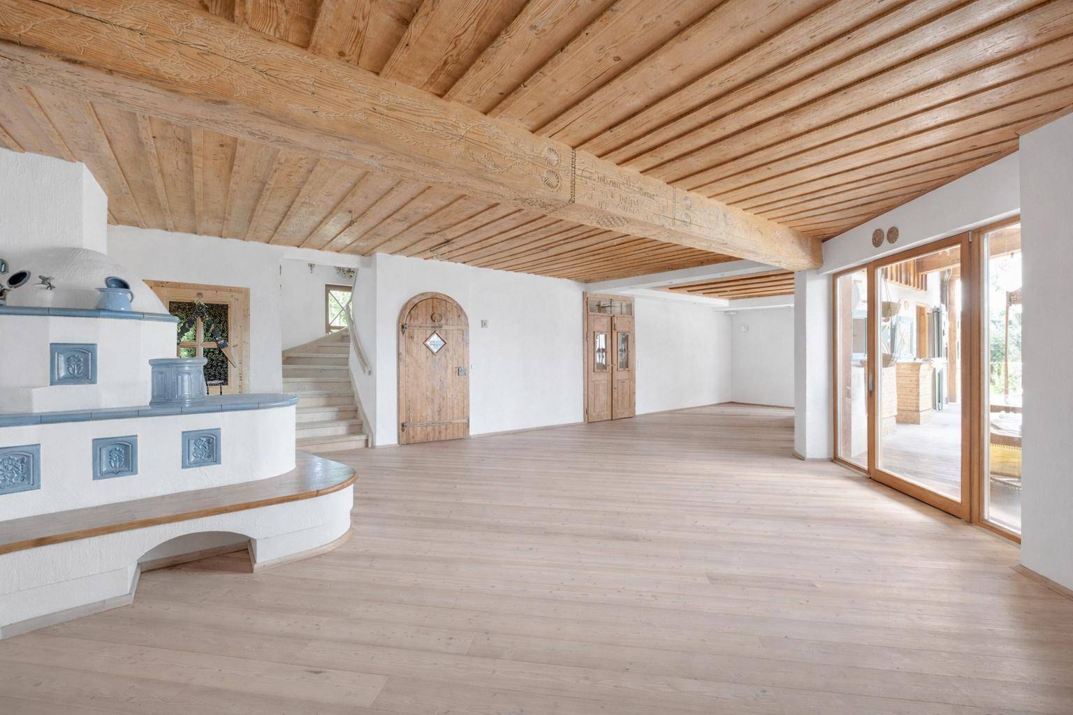 Heller Wohnbereich mit Holzbalkendecke, Kachelofen und Zugang zu einer Treppe und weiteren Räumen.