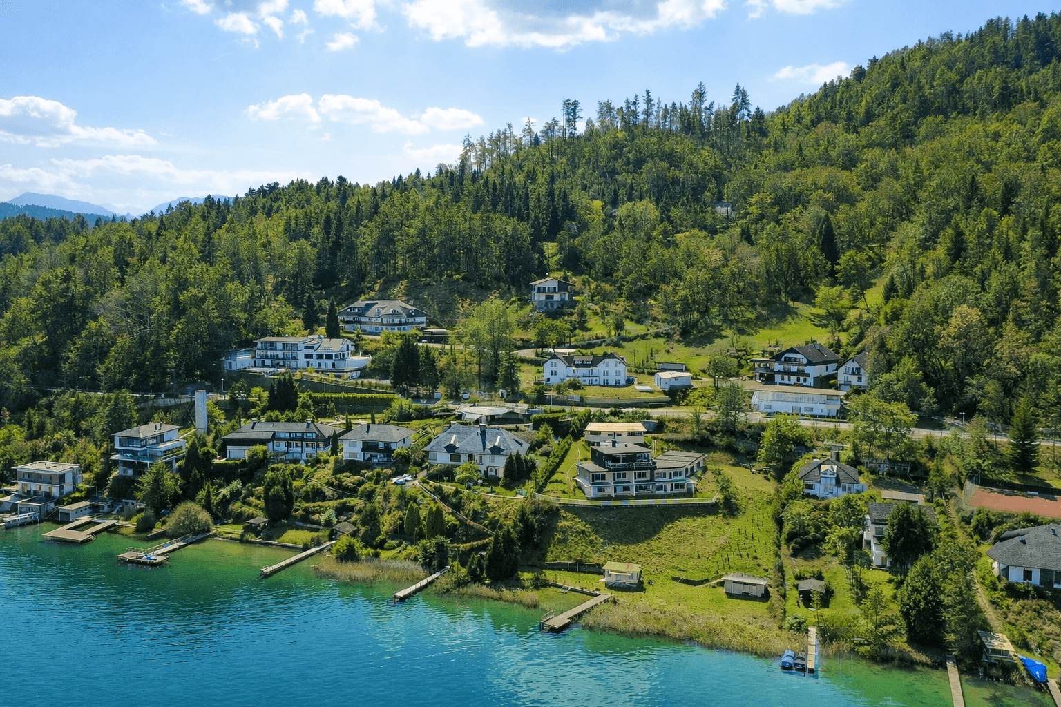 Luftaufnahme einer malerischen Siedlung am Seeufer, umgeben von grünen Hügeln und Wald.