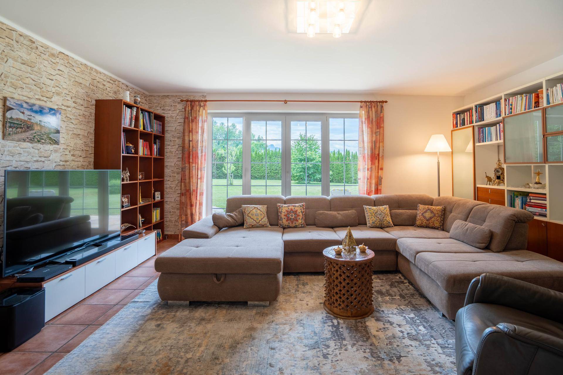 Gemütliches Wohnzimmer mit großer Eckcouch, Steinfliesenboden und Blick in den Garten.