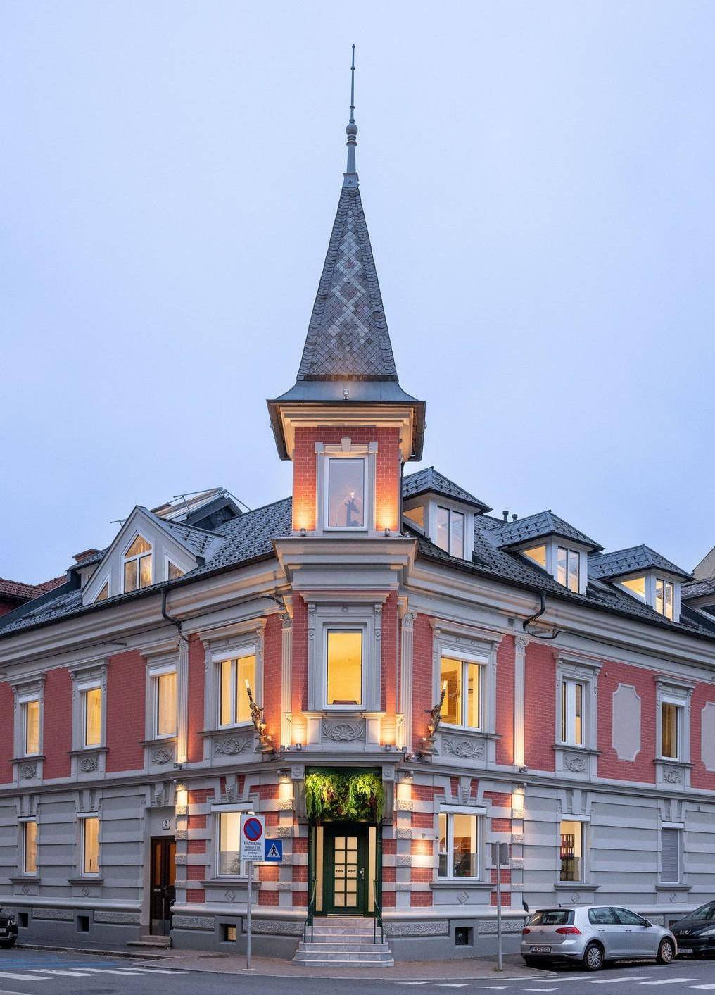Beleuchtete Fassade des historischen Eckgebäudes mit markantem Turm in der Abenddämmerung.