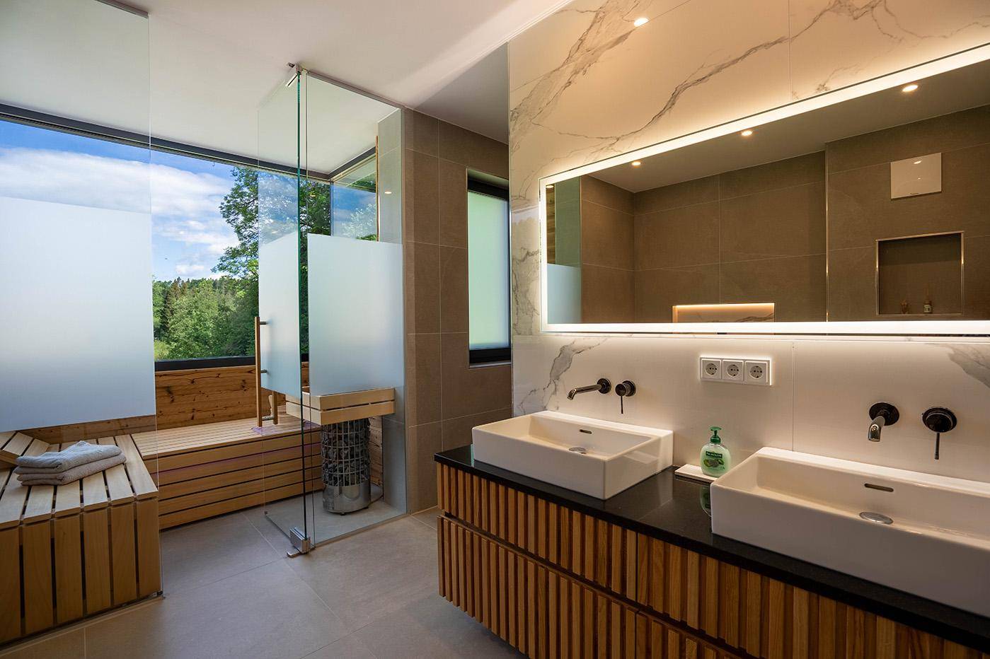 Luxuriöses Badezimmer mit Doppelwaschbecken, beleuchtetem Spiegel und integrierter Sauna mit Ausblick.