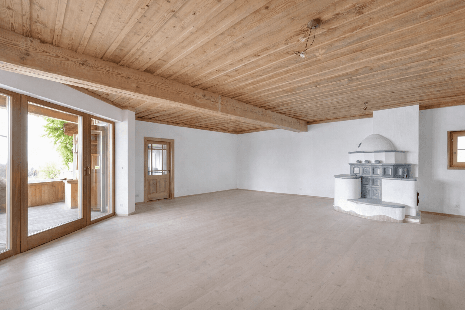 Geräumiges Wohnzimmer mit heller Holzbalkendecke, hellem Holzboden und einem traditionellen Kachelofen.