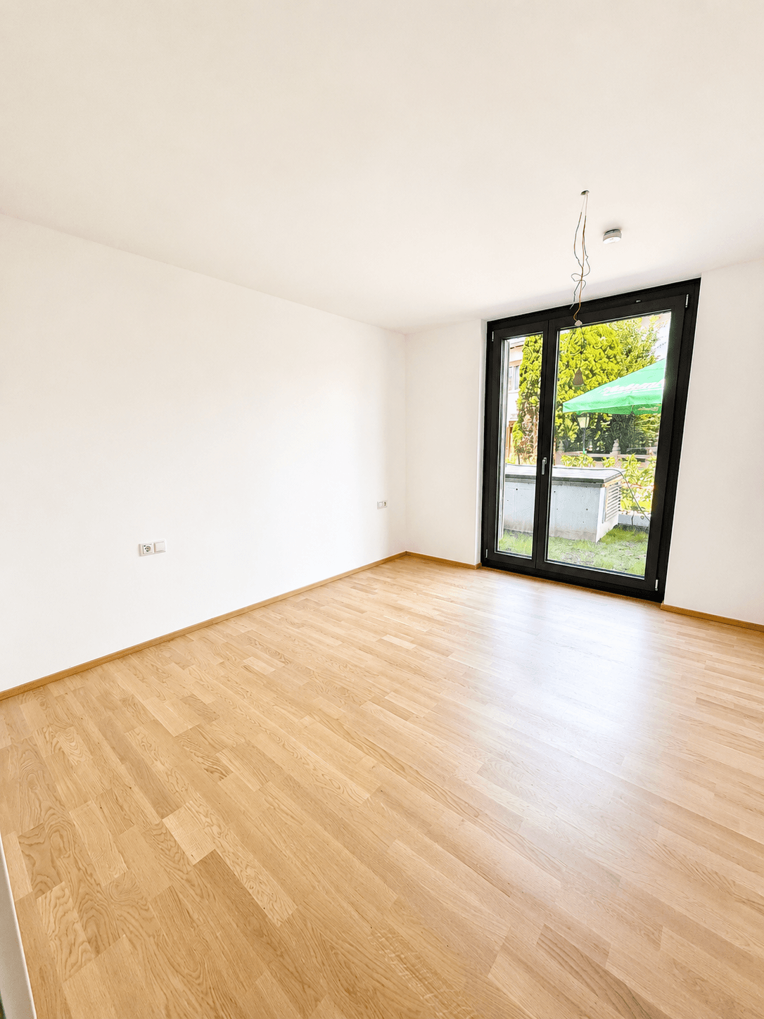 Heller, leerer Raum mit Holzboden und großem Fenster, das Zugang zu einem Gartenbereich bietet.