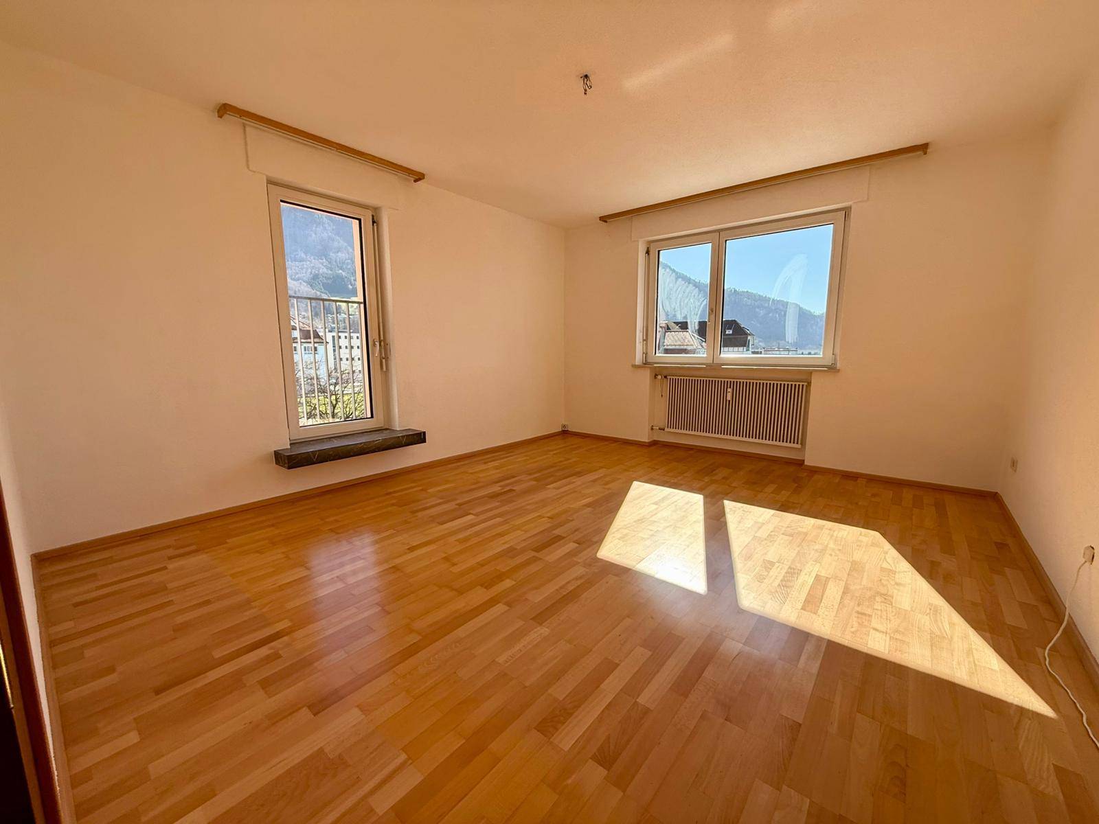 Sonniger Raum mit Parkettboden, großem Fenster und Blick auf die umliegende Landschaft.