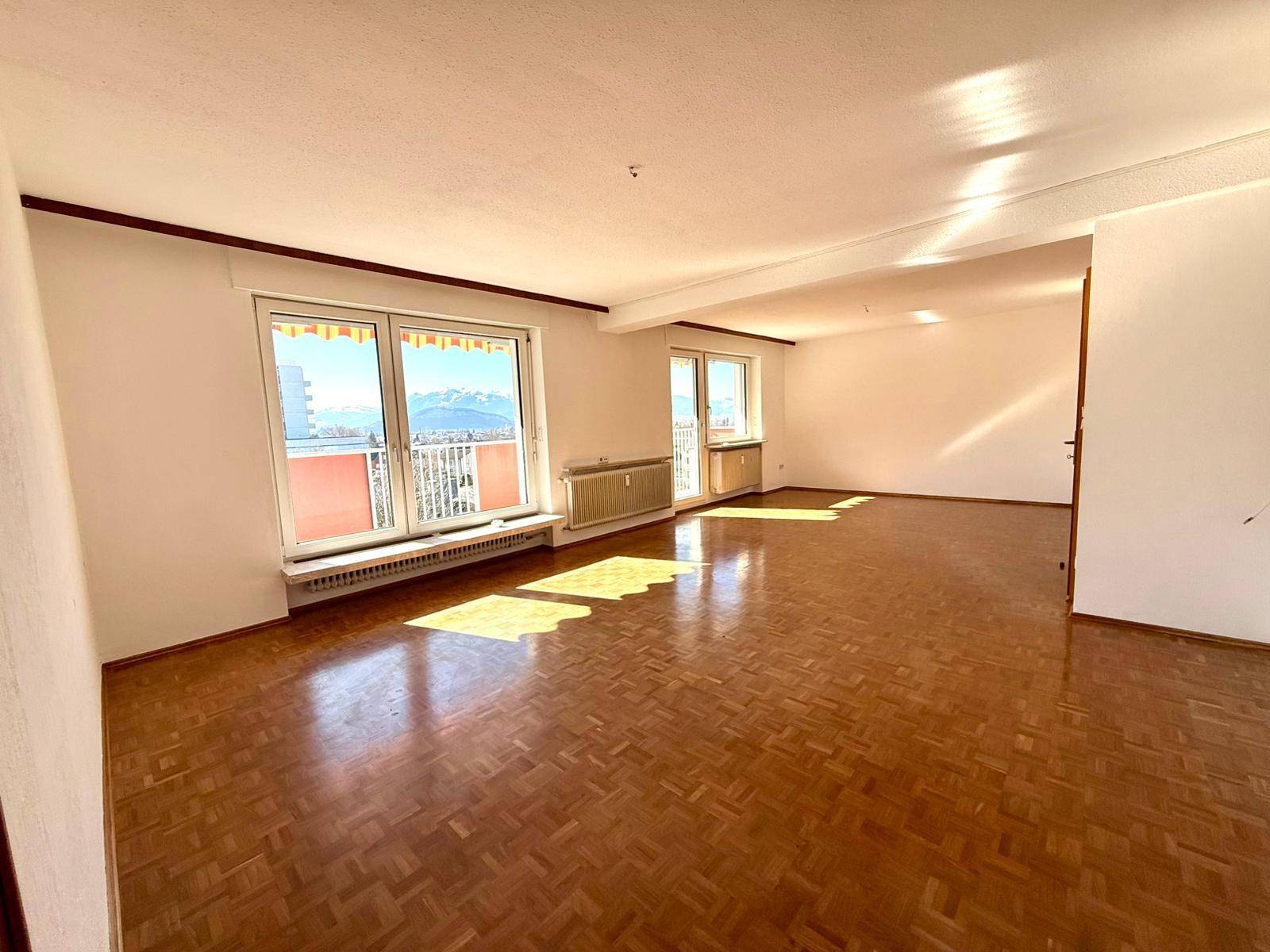 Geräumiges, leeres Wohnzimmer mit Parkettboden, großen Fenstern und viel natürlichem Licht.