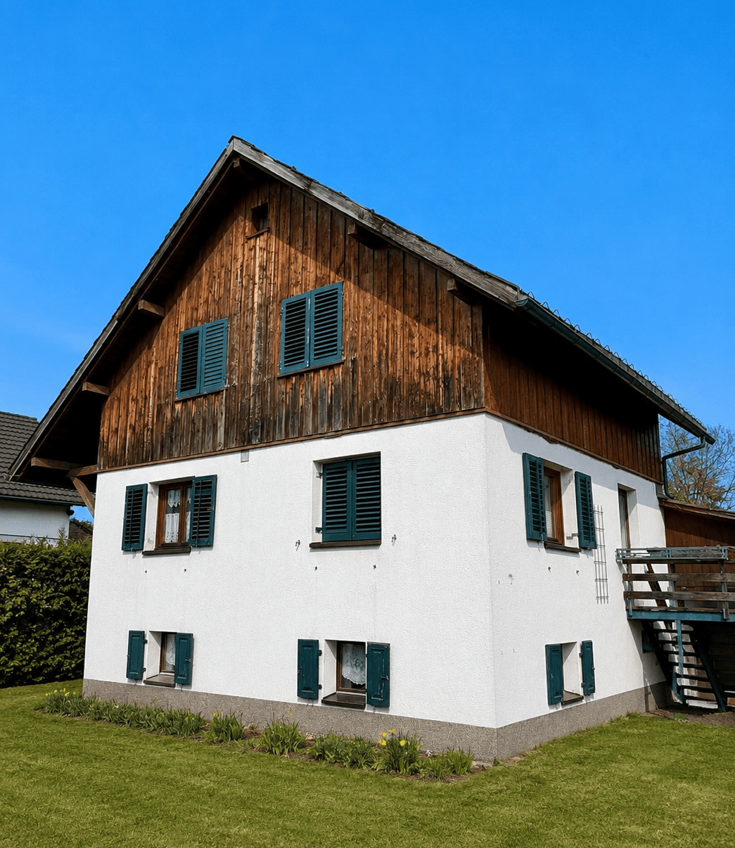 Freistehendes Haus mit weißer Fassade, Holzgiebel und grünen Fensterläden im Sonnenschein.