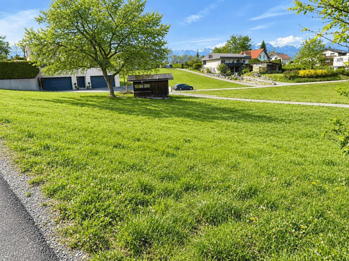 Sonnige Grünfläche mit einem kleinen Holzschuppen, umgeben von Wohnhäusern und Alpenpanorama.
