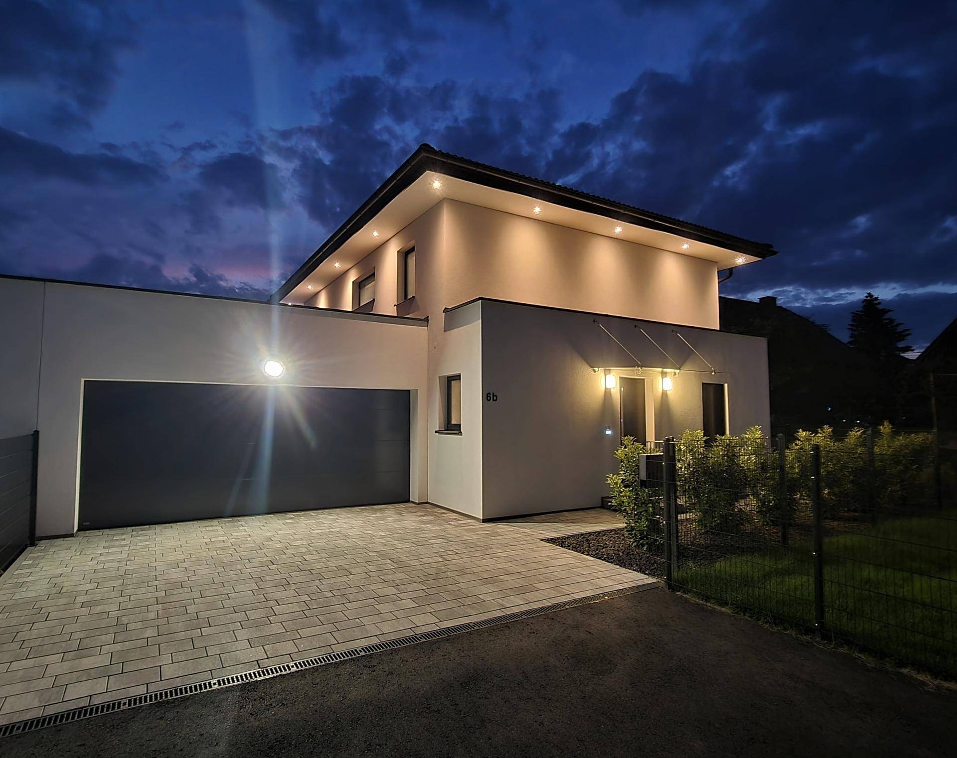 Moderne Hausfassade mit beleuchteter Garage und Eingangsbereich in der Abenddämmerung.