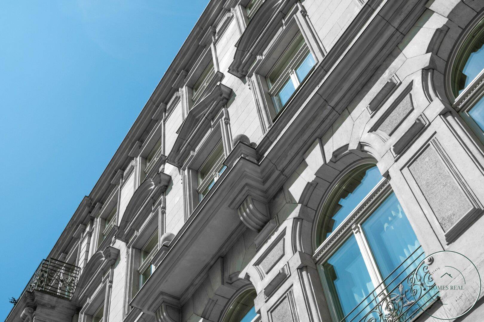Historische Fassade eines Gebäudes mit aufwendigen Verzierungen und großen Fenstern unter blauem Himmel.