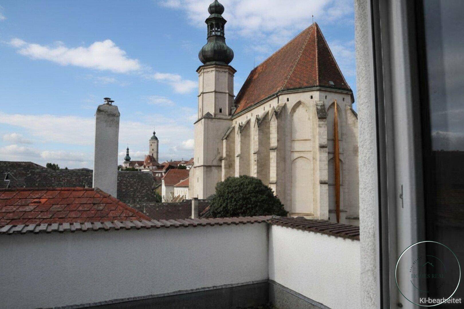 Blick von einer Terrasse auf eine historische Kirche und die Dächer einer Altstadt unter bewölktem Himmel.