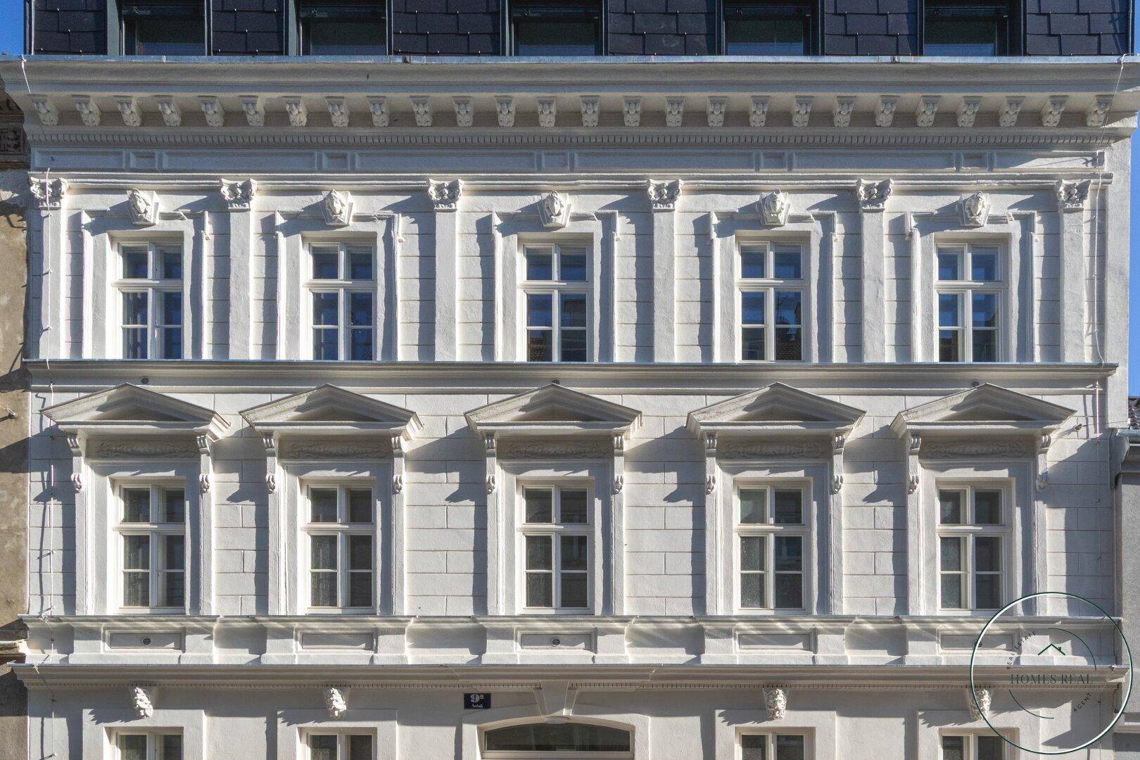 Historische Fassade eines Altbaus mit reich verzierten Fenstern und Stuckelementen unter einem Schieferdach.