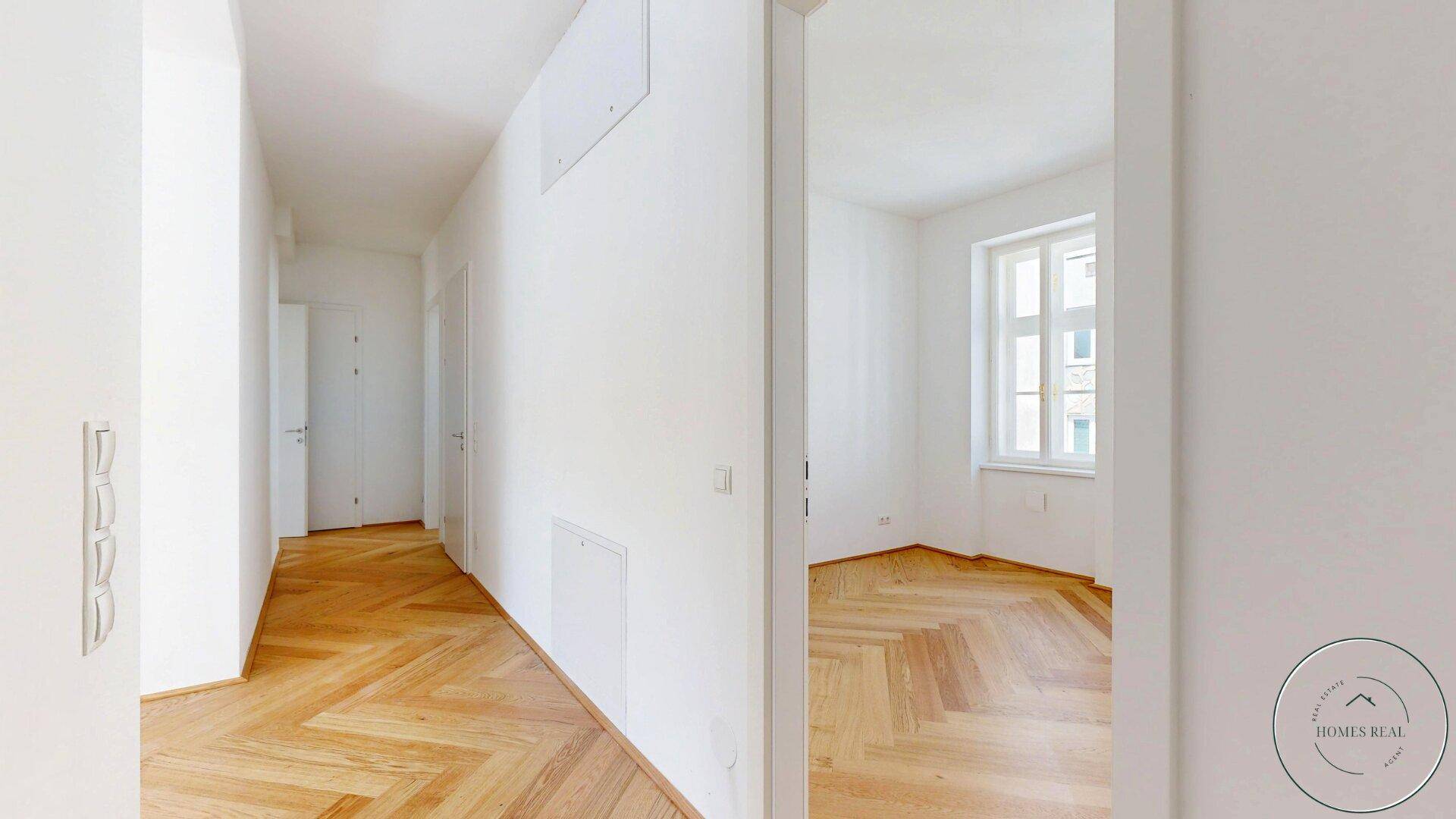 Heller Flur mit Fischgrätparkett und Blick in ein angrenzendes Zimmer mit Fenster.