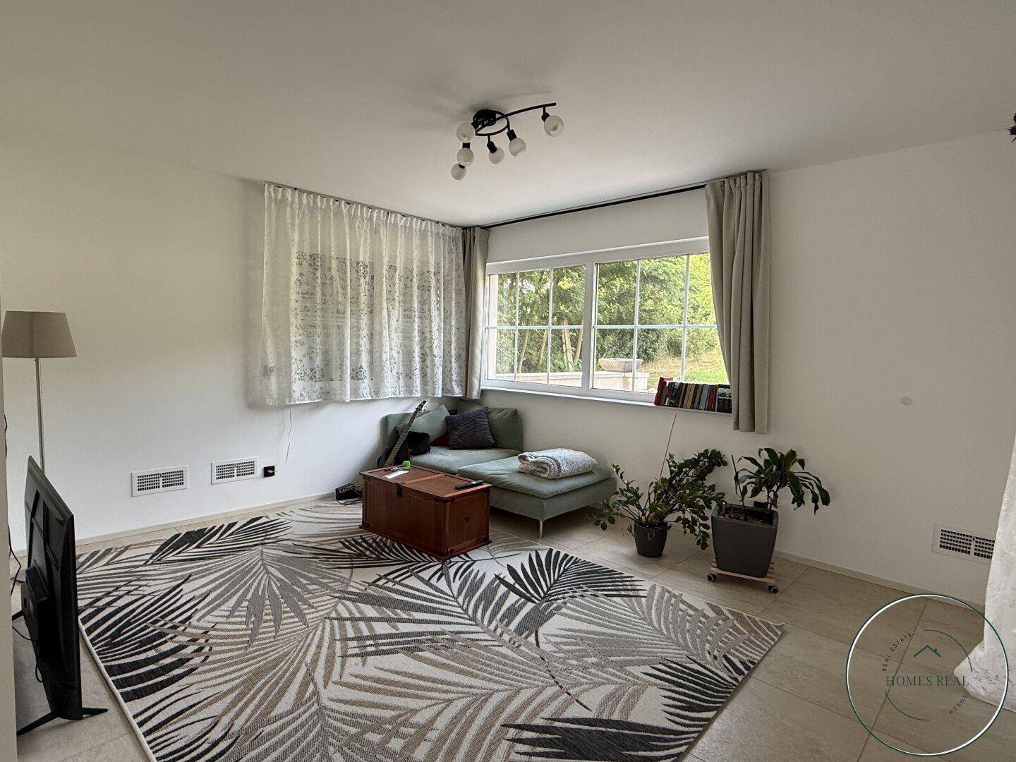 Gemütliches Wohnzimmer mit bequemer Couch, großem Teppich und Blick ins Grüne durch ein breites Fenster.