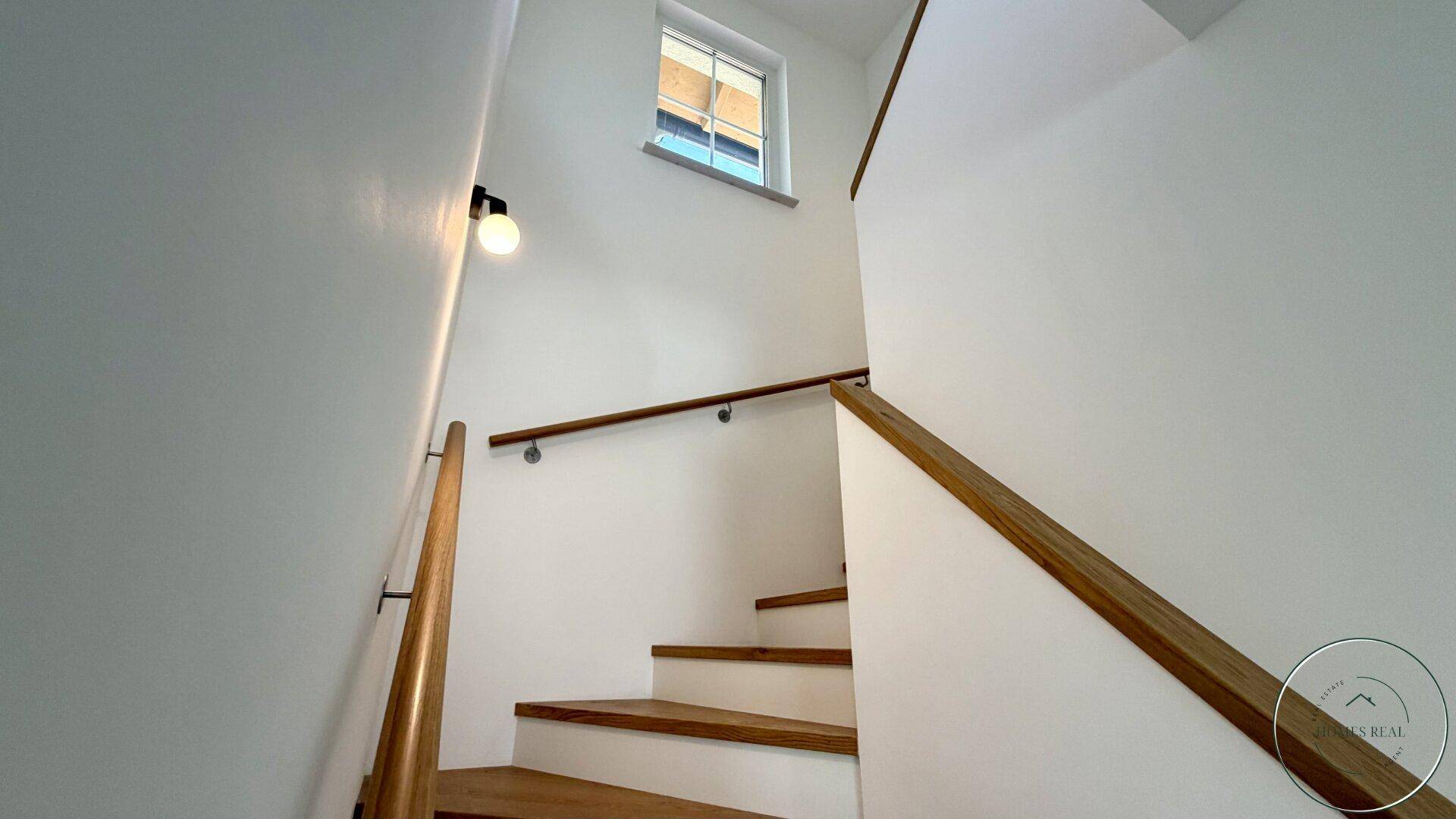 Heller Treppenaufgang mit Holzstufen und passendem Geländer, beleuchtet durch ein kleines Fenster.