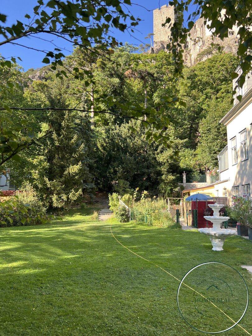 Weitläufiger Garten mit Rasen, Bäumen und einem Brunnen im Hintergrund.