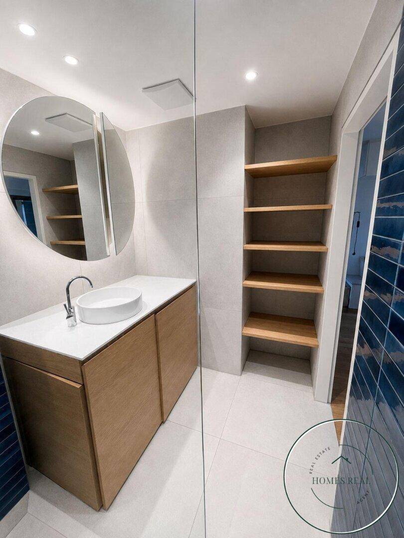 Modernes Badezimmer mit stilvollem Waschtisch, rundem Spiegel und integrierten Regalen.