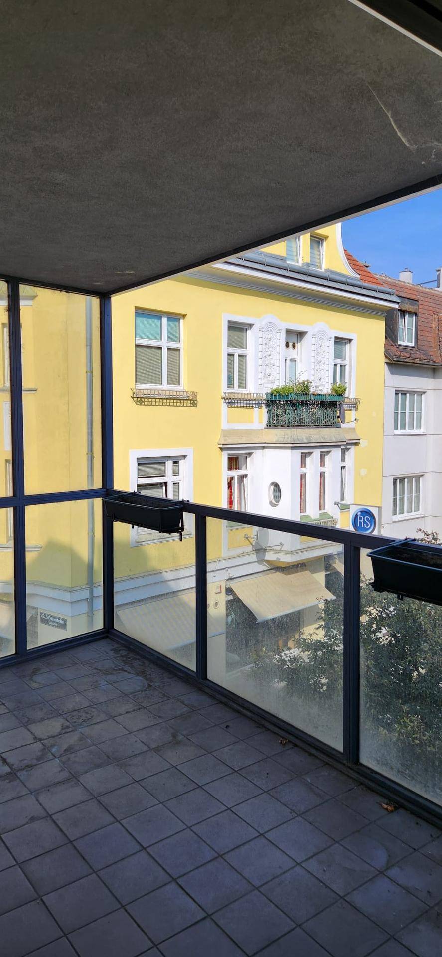 Balkon mit Fliesenboden und Glasgeländer, bietet einen Blick auf ein gegenüberliegendes Gebäude.