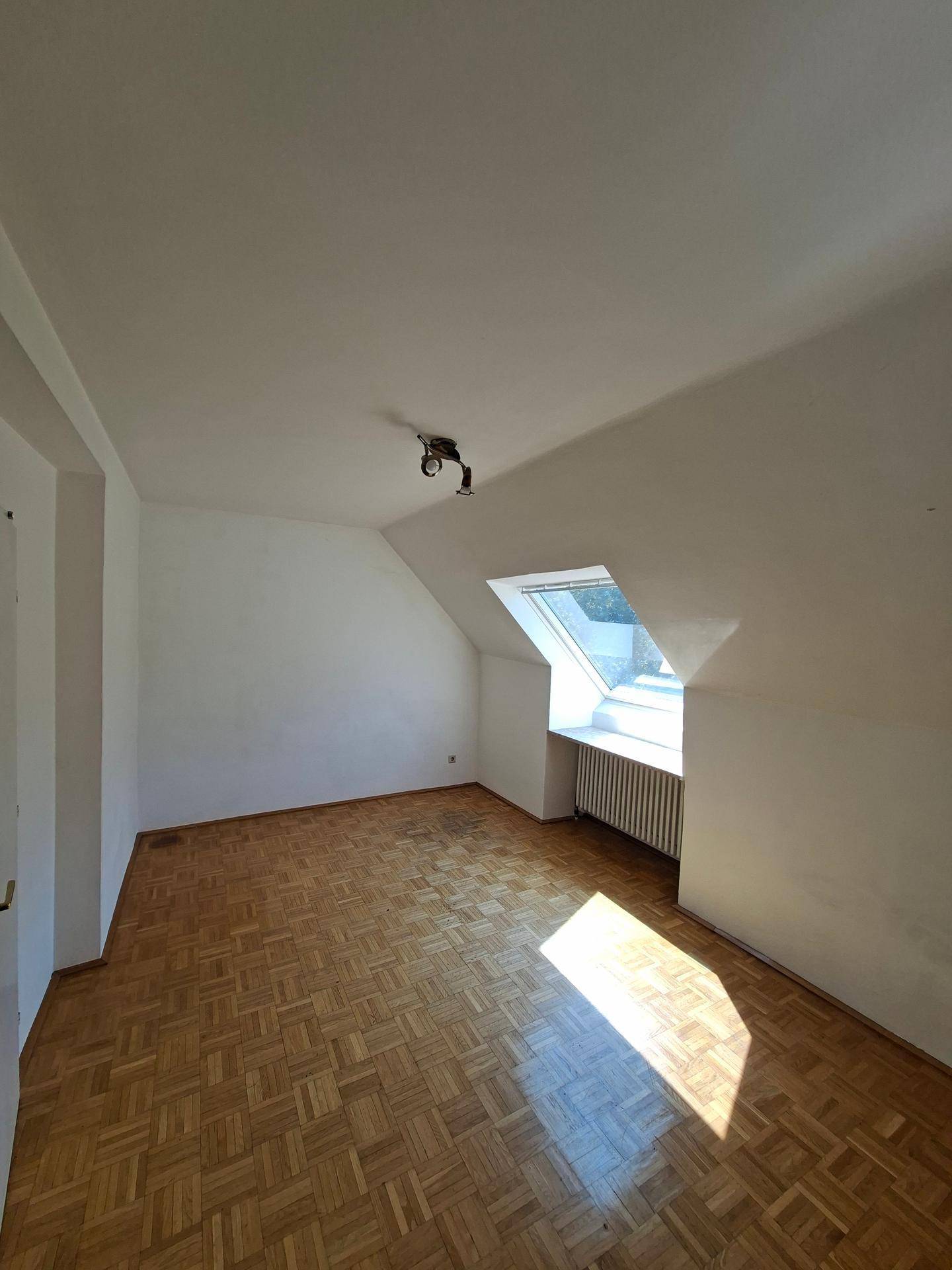 Helles Dachzimmer mit Parkettboden, schrägen Wänden und einem Dachfenster.