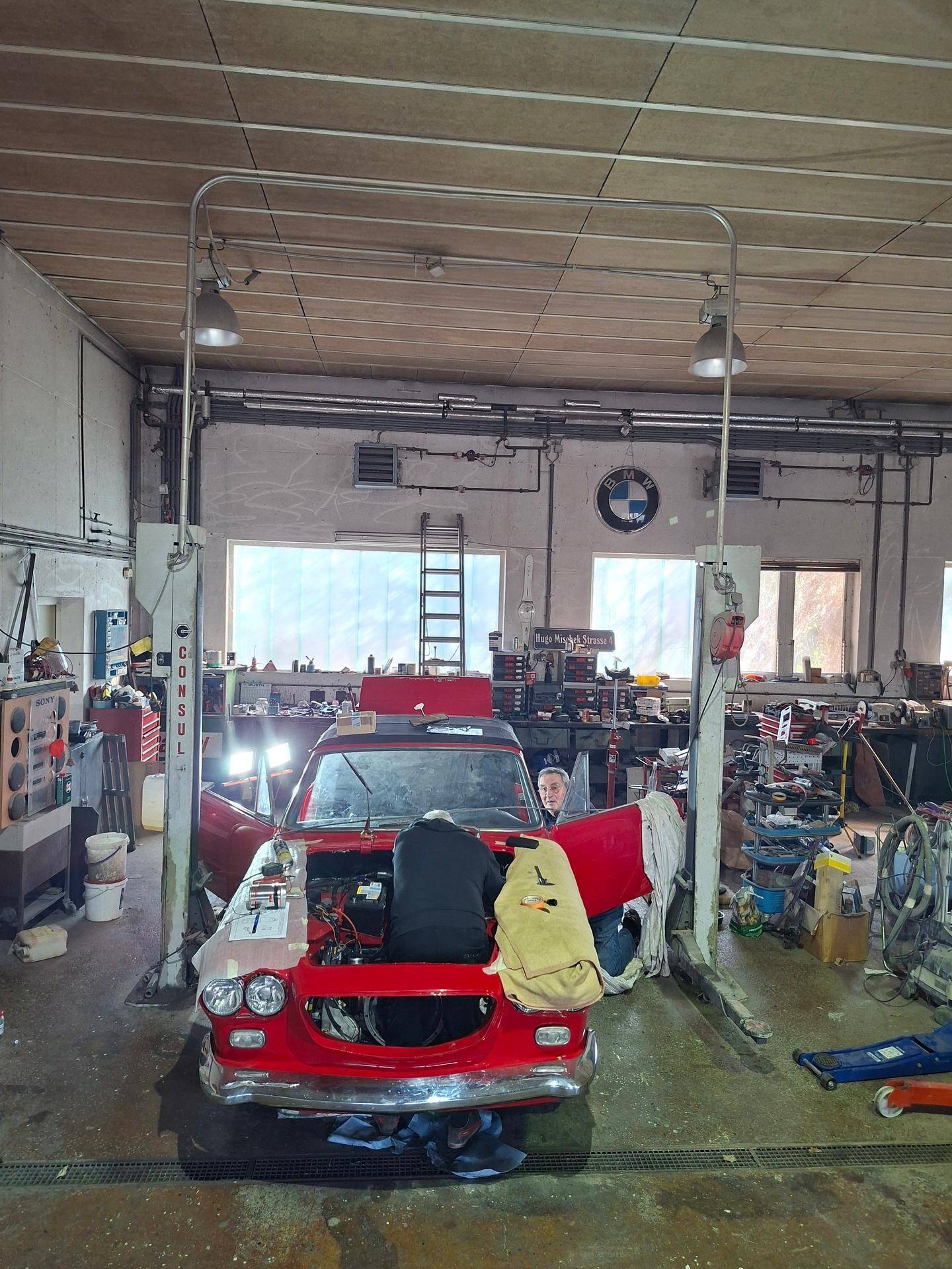 Detailansicht eines roten Oldtimers auf einer Hebebühne, während Mechaniker Reparaturen durchführen.
