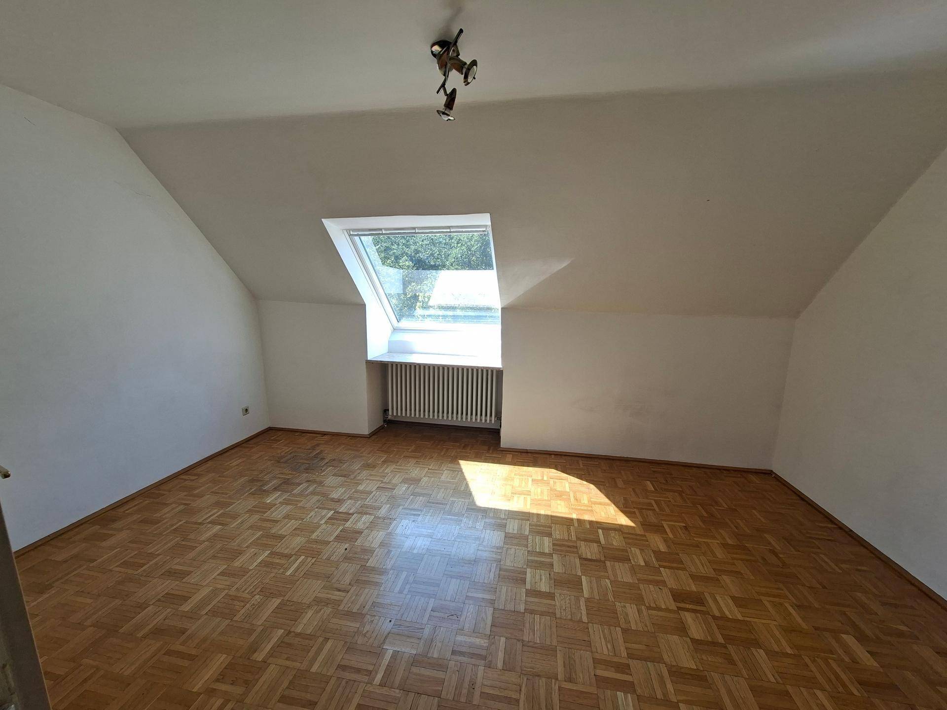 Sonniges Dachzimmer mit Parkettboden, Dachfenster und Deckenleuchte.