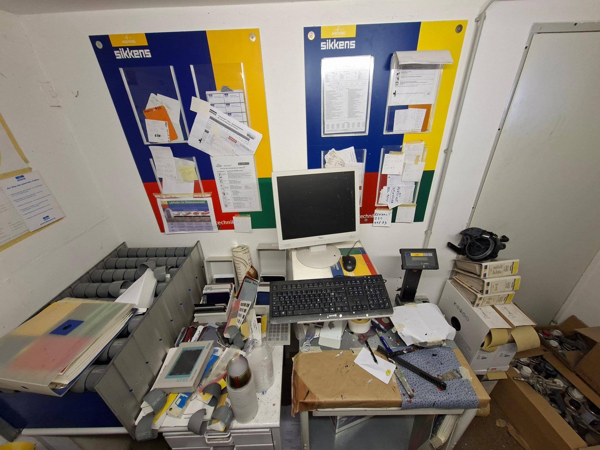 Detailansicht eines Arbeitstisches in einem Farbmischraum mit Computer und Dokumenten.