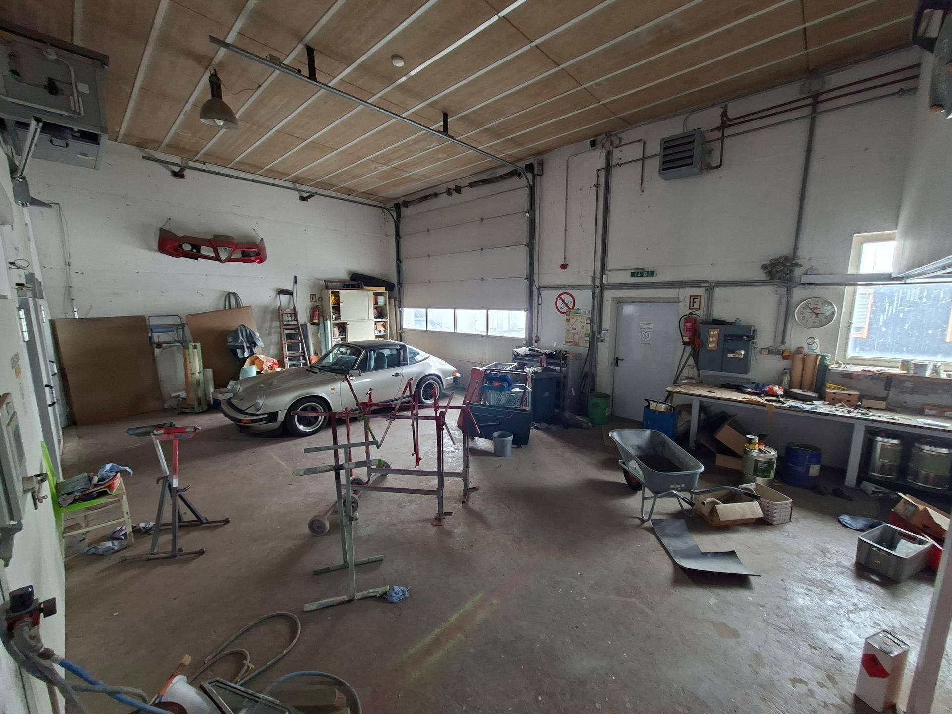 Weitläufige Werkstatt mit einem silbernen Porsche, einer Lackierkabine und Werkbänken entlang der Wand.