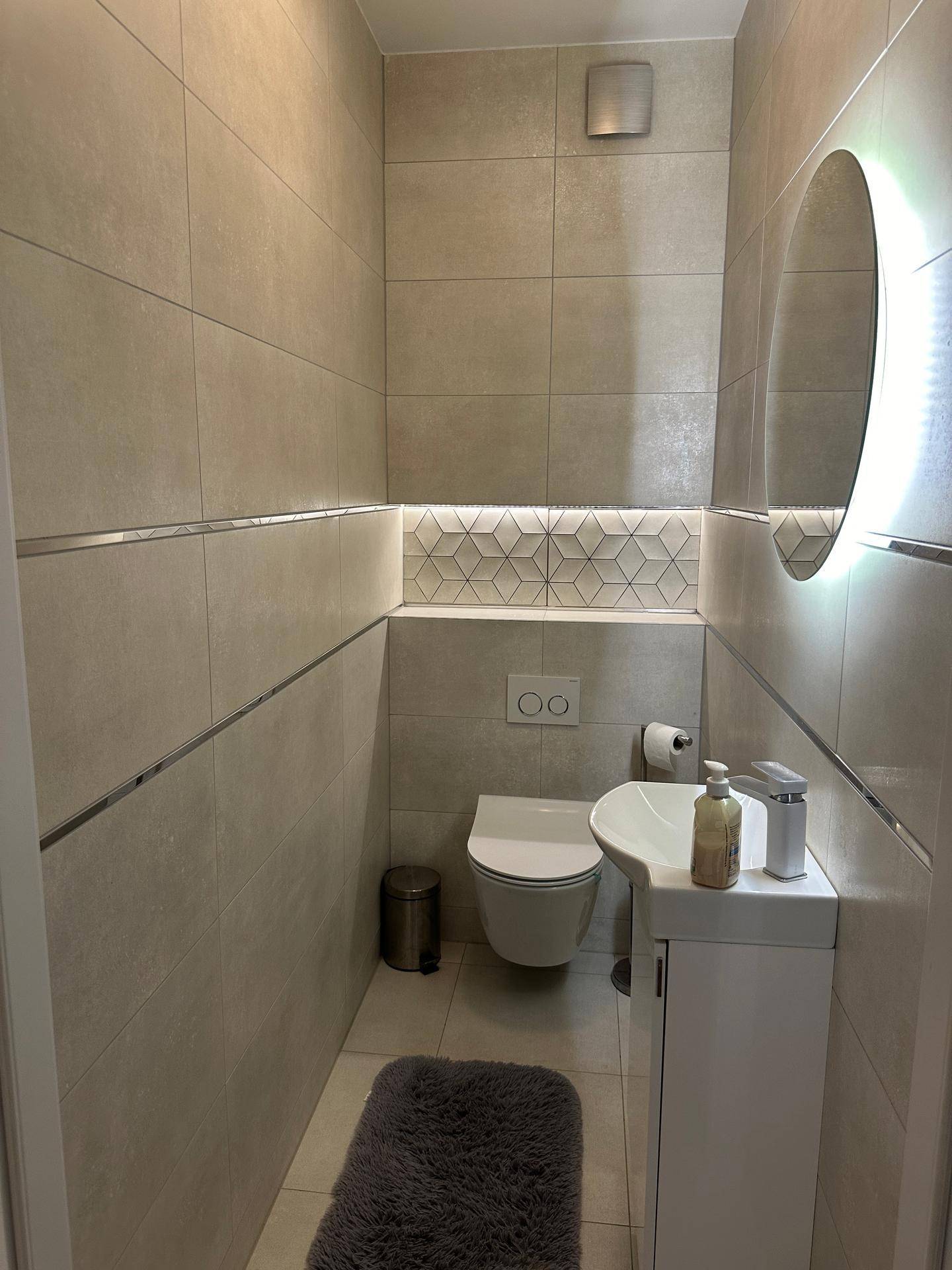 Modernes Gäste-WC mit Wand-WC, kleinem Waschbecken und beleuchtetem Spiegel.