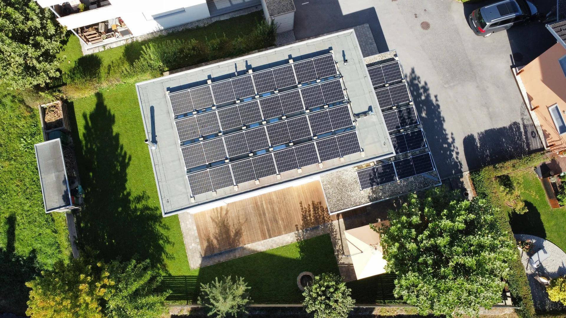 Luftaufnahme eines Hauses mit Photovoltaikanlage auf dem Flachdach und umliegendem Garten.