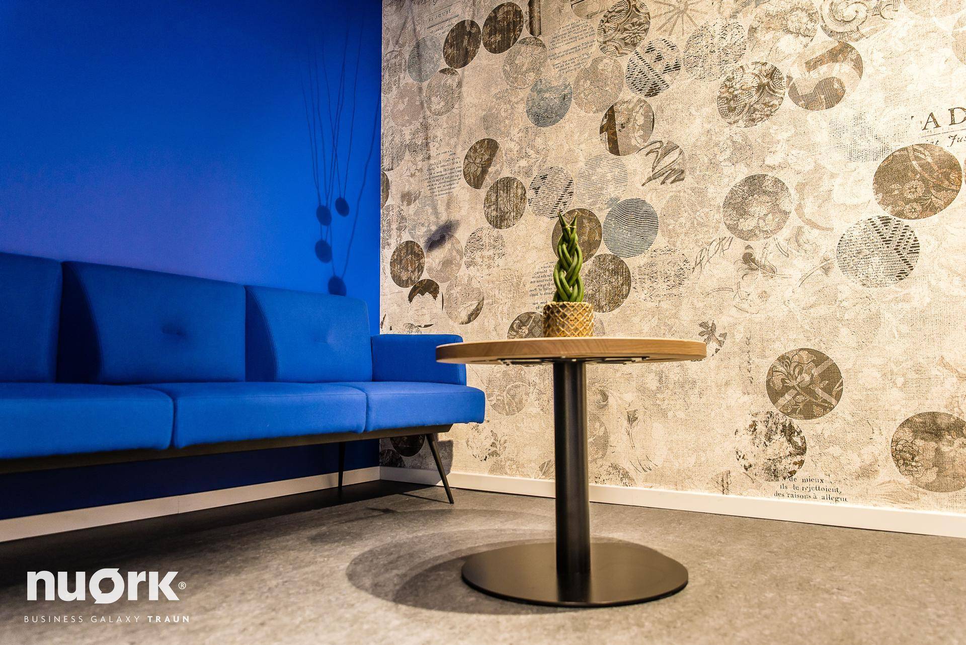 Gemütlicher Lounge-Bereich im Flur mit blauen Sesseln, einem kleinen Tisch und Blick in einen Büroraum.
