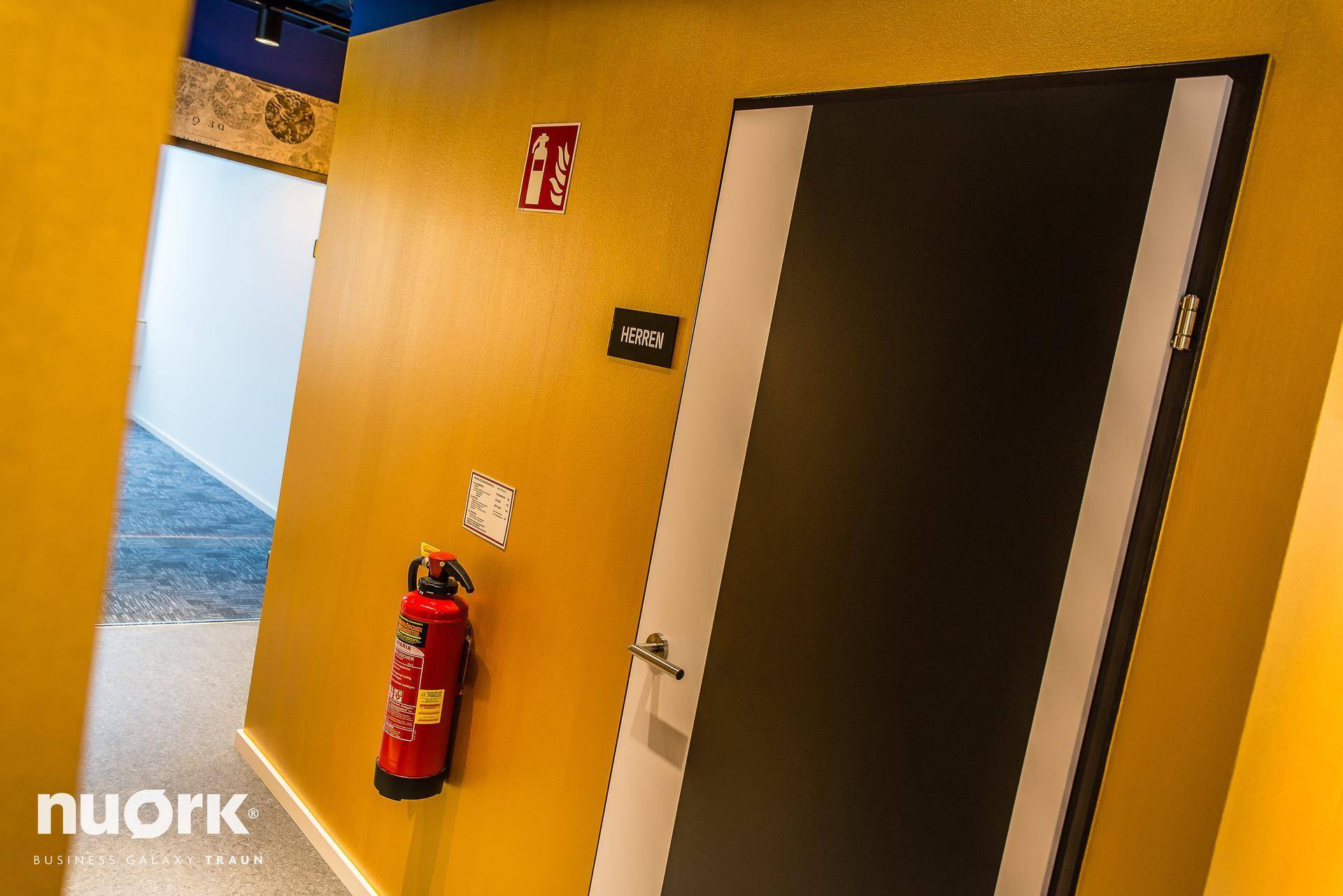 Flur mit gelben Wänden, einem Feuerlöscher und einer Tür, die zu den Herren-Toiletten führt, in einem Bürogebäude.