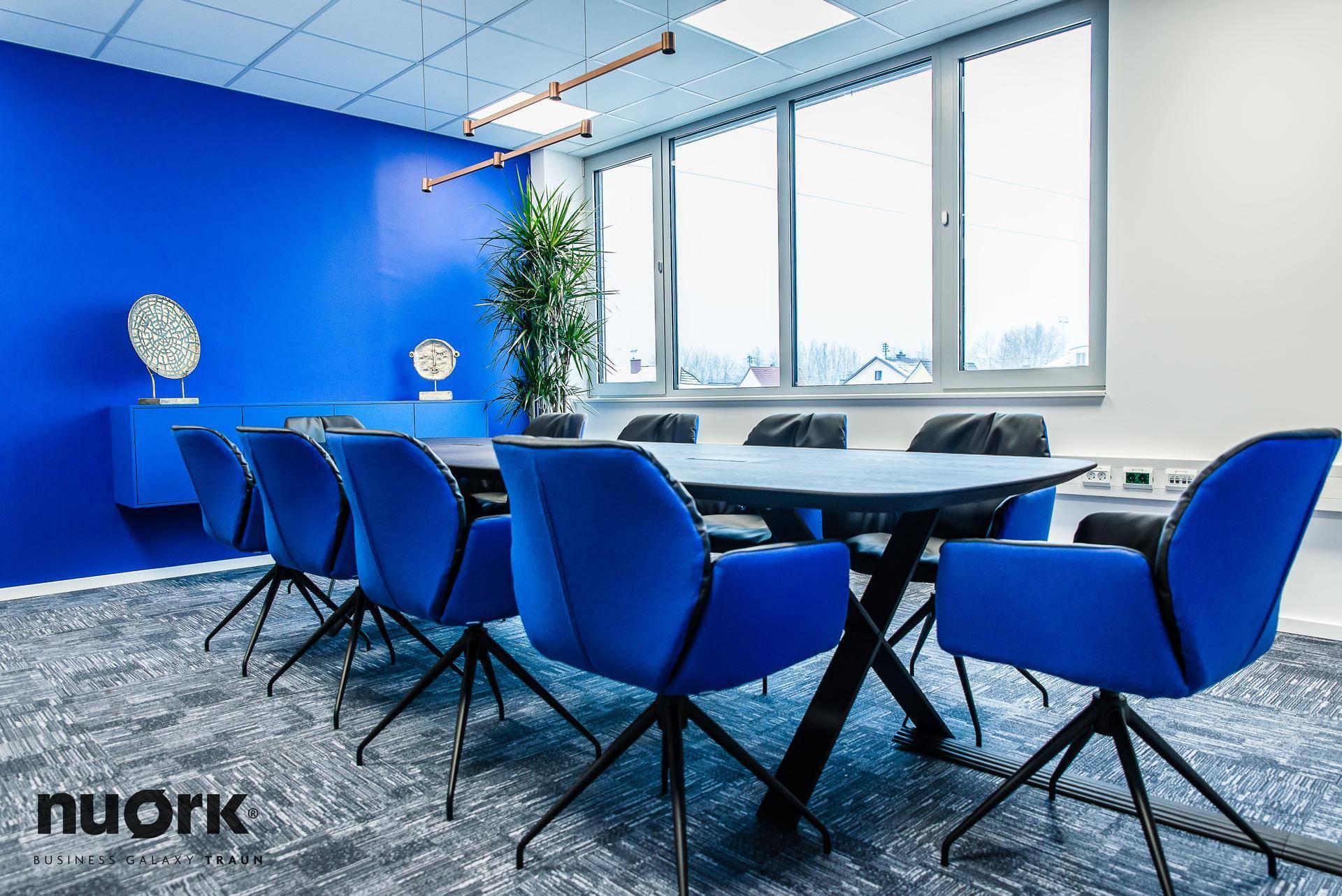 Moderner Konferenzraum mit großem Bildschirm, blauen Stühlen und Blick aus dem Fenster auf die Umgebung.