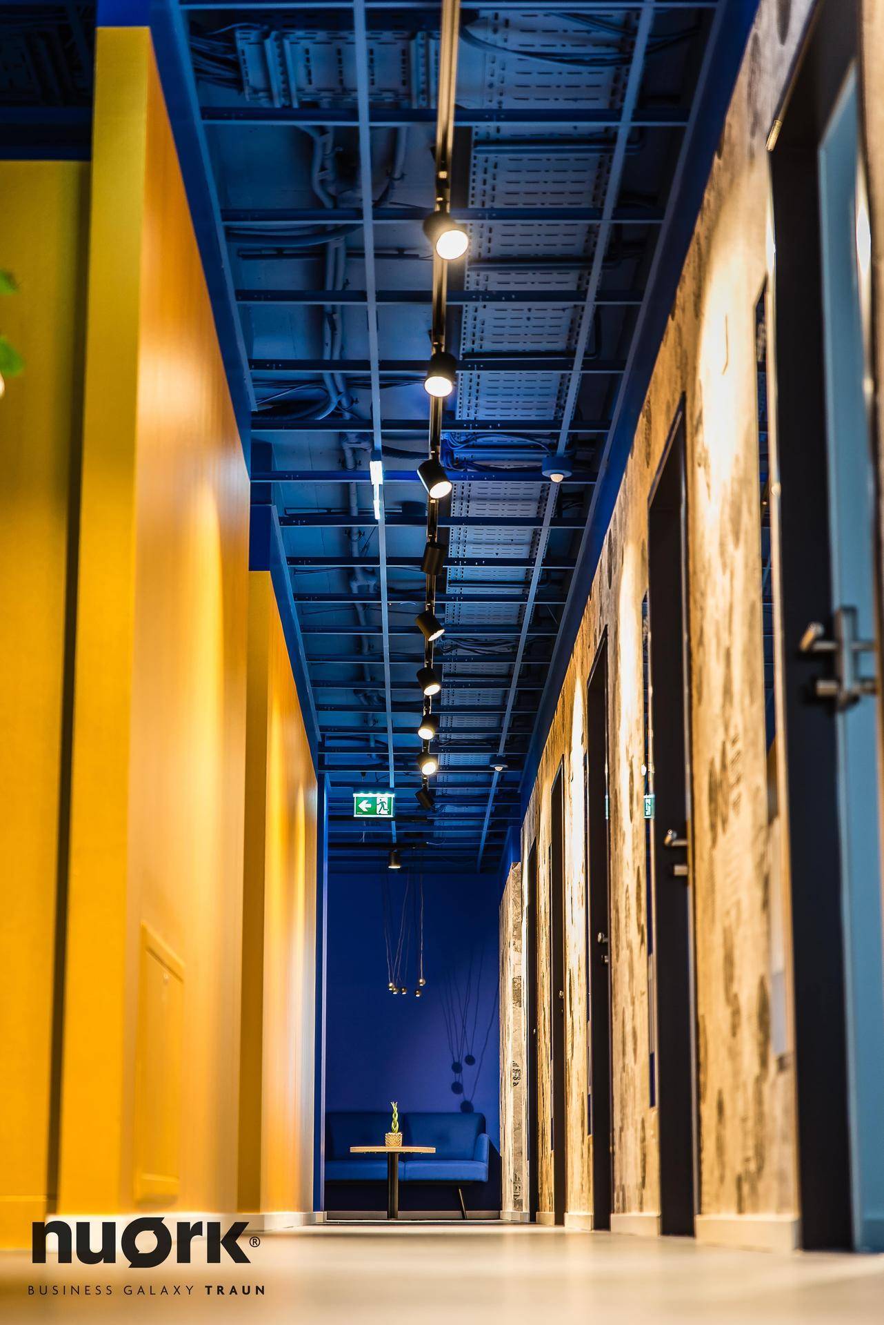Langer Flur mit blauer Decke, gelben Wänden und sichtbaren Installationen, die einen industriellen Charme verleihen.