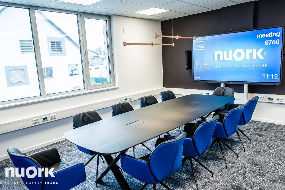 Moderner Konferenzraum mit großem Tisch, blauen Stühlen und einem großen Bildschirm, der viel natürliches Licht erhält.