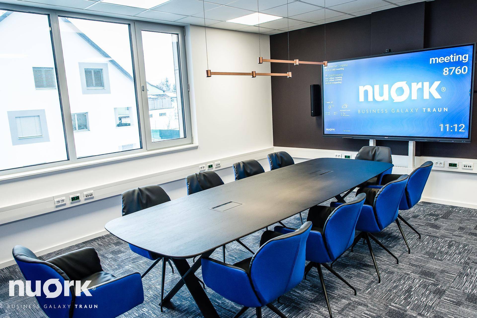 Heller Konferenzraum mit großem Tisch, blauen Stühlen und Blick auf die Umgebung, der eine inspirierende Atmosphäre schafft.